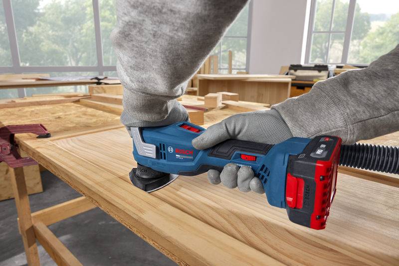 Eine Person in Handschuhen verwendet ein kabelloses Elektroschleifergerät, um eine Holzoberfläche in einer Werkstatt zu schleifen, in die Licht durch große Fenster strömt.