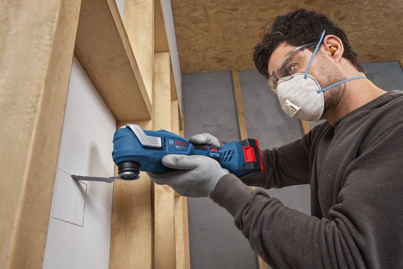 Eine Person, die Schutzbrille und Maske trägt, benutzt ein Elektrowerkzeug, um ein Quadrat in eine weiße Wand zu schneiden, die in einen Holzrahmen eingelasst ist.