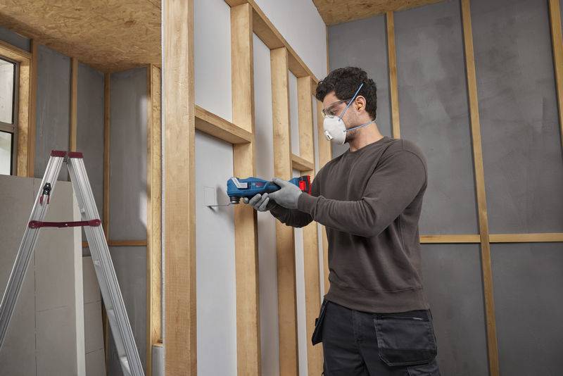 Ein Mann in Schutzausrüstung benutzt ein Werkzeug an einem Holzrahmen in einem unfertigen Raum mit einer Stehleiter in der Nähe, was auf Bauarbeiten hinweist.