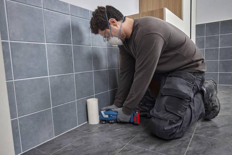 Ein Arbeiter kniet auf einem Fliesenboden und trägt mit einem Werkzeug Fugenmörtel auf die Wandfliesen auf. Er trägt Schutzbrille, Maske und Handschuhe.