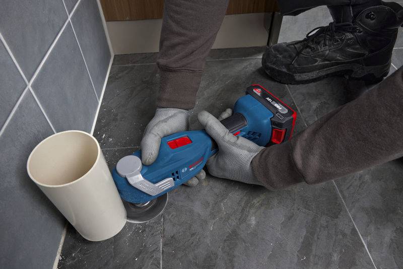 Eine Person mit Handschuhen benutzt ein blaues Elektrowerkzeug, um eine Fliese in der Nähe eines Rohrs auf einem gefliesten Boden zu schneiden.