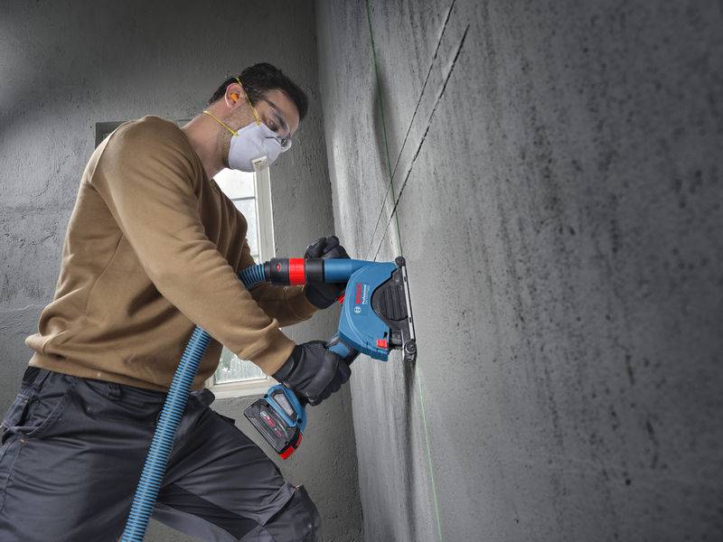 Eine Person in Schutzausrüstung verwendet ein Elektrowerkzeug an einer Betonwand und konzentriert sich auf die präzise Ausrichtung von Linien. Sonnenlicht fällt durch ein nahegelegenes Fenster ein.