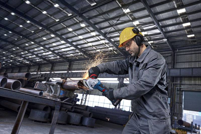 Ein Arbeiter in Schutzausrüstung verwendet einen Winkelschleifer an einem Metallrohr in einem Industrielager und erzeugt dabei Funken.