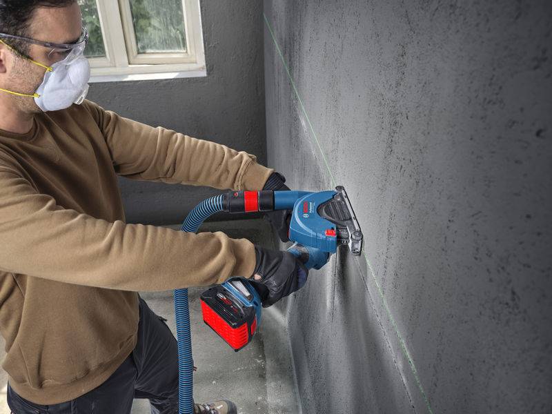 Eine Person trägt Schutzausrüstung, einschließlich einer Maske und Schutzbrille, und verwendet ein Elektrowerkzeug, um eine gerade Linie auf einer Betonwand in einem Innenraum zu schneiden.