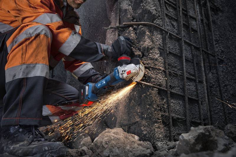 Ein Bauarbeiter in orangefarbener Schutzausrüstung benutzt ein Elektrowerkzeug, um Bewehrungsstahl an einer Betonwand zu schneiden, wobei helle Funken entstehen.