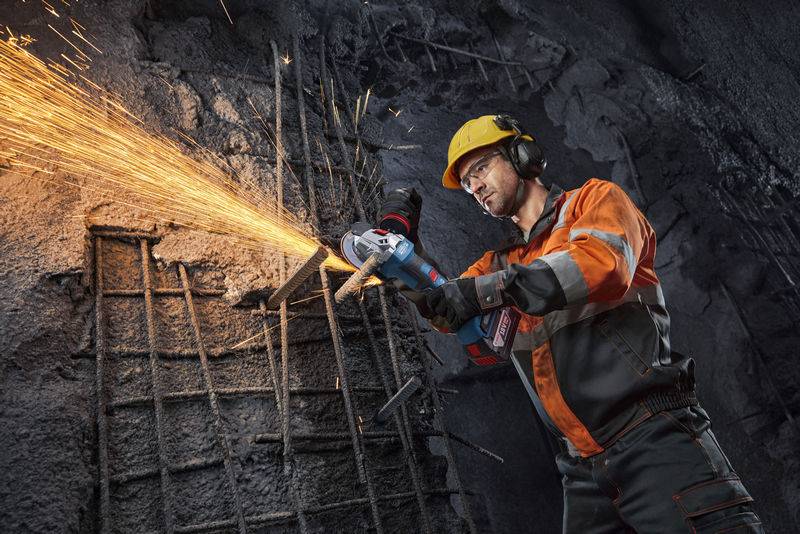 Ein Bauarbeiter in Schutzausrüstung benutzt ein Elektrowerkzeug, um Metallstäbe zu schneiden, wobei Funken gegen eine Betonwand in einer schwach beleuchteten Umgebung entstehen.