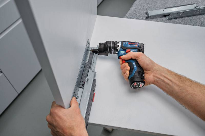 Eine Person verwendet einen kabellosen Bohrer, um eine Metallschiene an der Seite eines weißen Schrankpanels zu befestigen und bereitet damit die Möbelmontage vor.