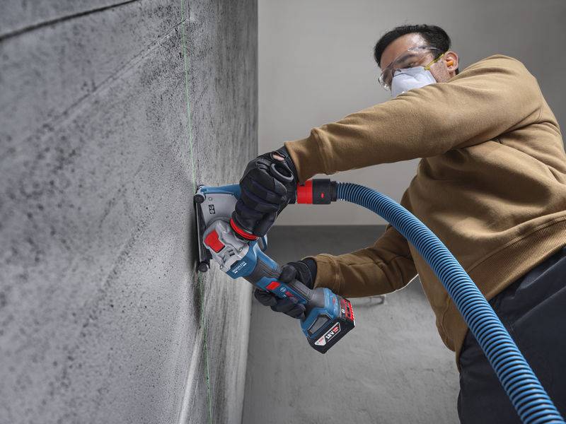 Eine Person in Schutzausrüstung verwendet ein Elektrowerkzeug, um eine Linie in eine Betonwand in einem Innenraum zu schneiden.