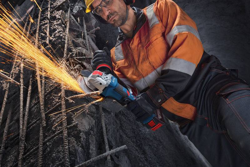 Ein Bauarbeiter in Schutzausrüstung verwendet einen Winkelschleifer, um Metallbewehrung zu schneiden, wodurch Funken in einem schwach beleuchteten Bereich herumfliegen.