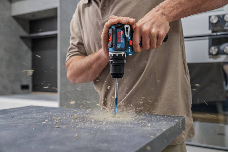 Eine Person bohrt mit einer Bosch-Akkubohrmaschine in eine Oberfläche, wobei sich Holzstaub verteilt.