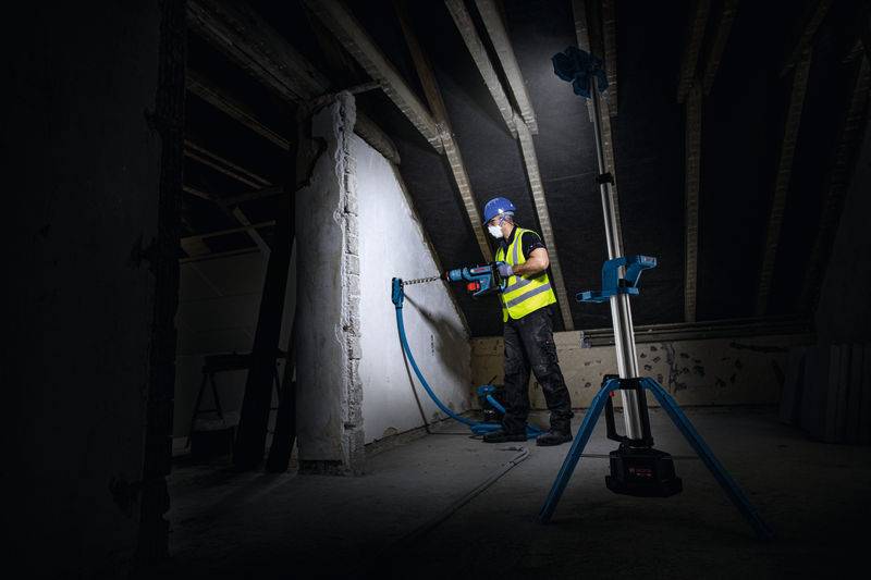 Ein Bauarbeiter in einer leuchtend farbigen Warnweste und mit Schutzhelm nutzt ein Elektrowerkzeug, um in einem dunkel beleuchteten Dachboden in eine Wand zu bohren.