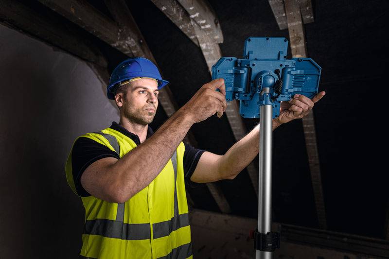 Ein Bauarbeiter mit blauem Helm und gelbem Sicherheitsweste stellt ein blaues Panel auf einem Metallständer ein, in einem schwach beleuchteten Raum mit Holzbalken.