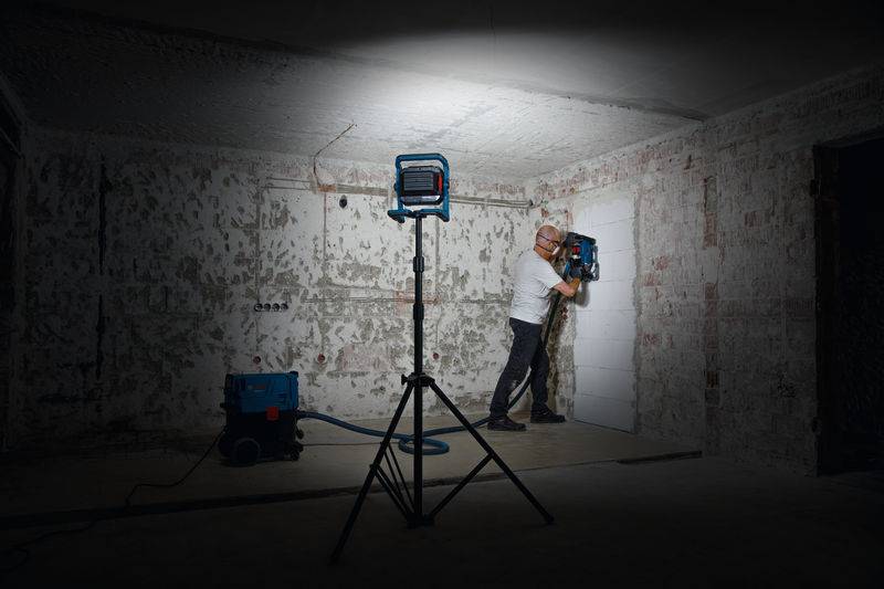 Eine Person mit Schutzhelm und Schutzausrüstung benutzt ein Elektrowerkzeug an einer Wand in einem schwach beleuchteten, unvollendeten Raum. Eine Arbeitsleuchte erhellt den Bereich.