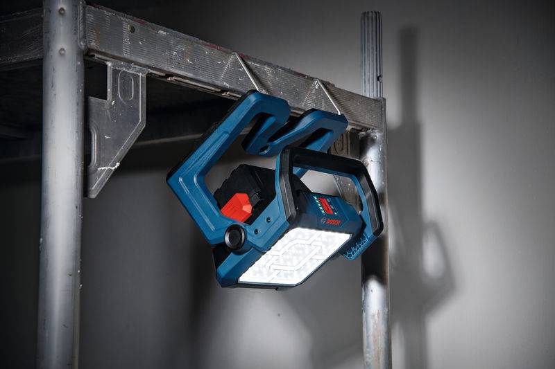 Eine tragbare LED-Arbeitsleuchte mit blauem Rahmen hängt an einem Metallgerüst und beleuchtet eine düstere Baustelle mit einstellbarer Helligkeit.