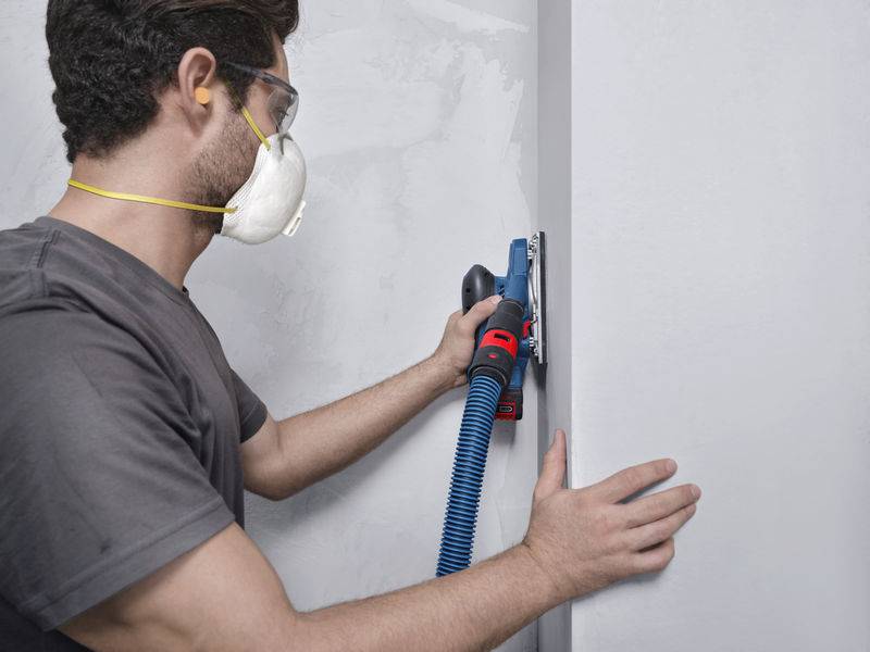 Eine Person, die eine Schutzbrille und eine Maske trägt, schleift eine Wand mit einem elektrischen Schleiferabtrag, wobei sie diesen mit beiden Händen hält.