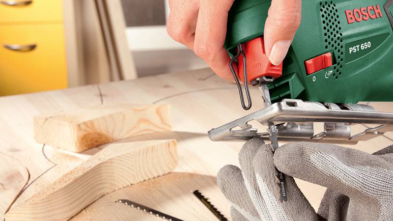 Eine Person nutzt eine Bosch PST 650 Stichsäge, um ein Holzstück auf einer Werkbank zu schneiden, wobei Schutzhandschuhe sichtbar sind.