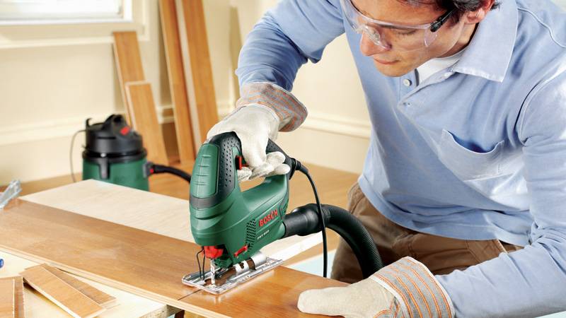 Eine Person benutzt eine grüne Stichsäge zum Schneiden von Holz auf einer Werkbank und trägt Schutzbrille und Handschuhe zum Schutz in einer Werkstattumgebung.