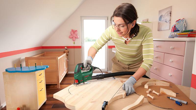 Eine Person in Schutzausrüstung benutzt ein Elektrowerkzeug, um Holz in einem Kinderzimmer zu schneiden, mit einem Gitterbett und Kindermöbeln im Hintergrund.
