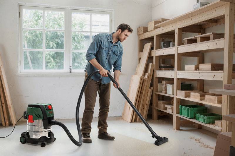 Eine Person benutzt einen Staubsauger in einer Werkstatt, umgeben von Holzregalen und Materialien, was auf eine laufende Reinigungsaufgabe hindeutet.