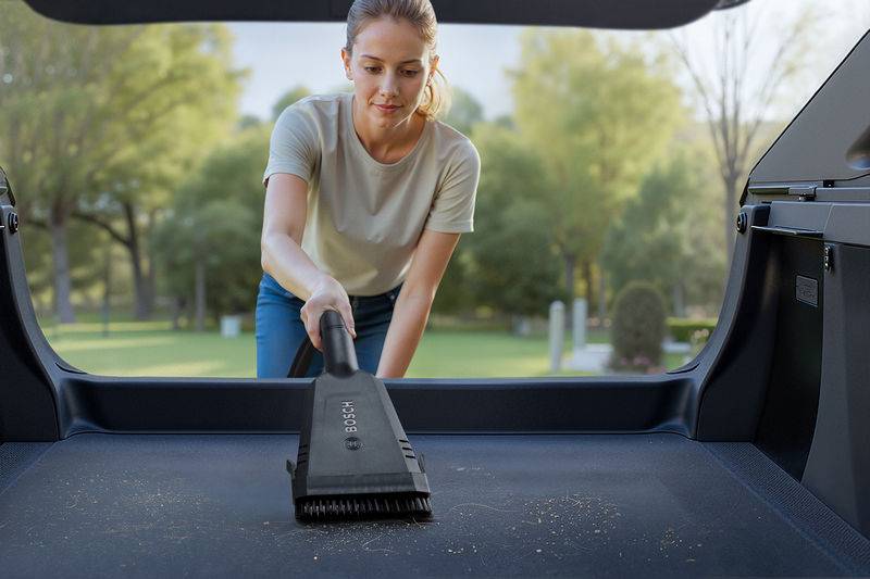 Eine Person benutzt einen Autostaubsauger, um Verschmutzungen aus dem Kofferraum eines Fahrzeugs an einem sonnigen Ort im Park zu entfernen.