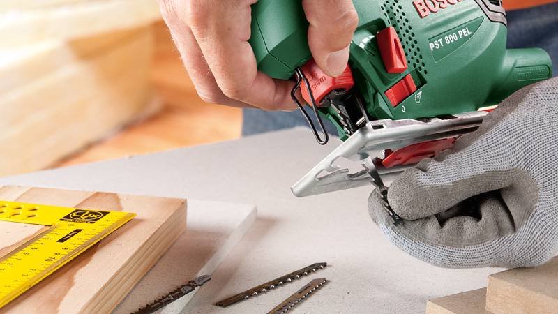 Eine Person, die eine grüne Stichsäge zum Schneiden von Holz benutzt, trägt Schutzhandschuhe. Ein gelbes Lineal und Ersatzklingen liegen in der Nähe auf einem Tisch.