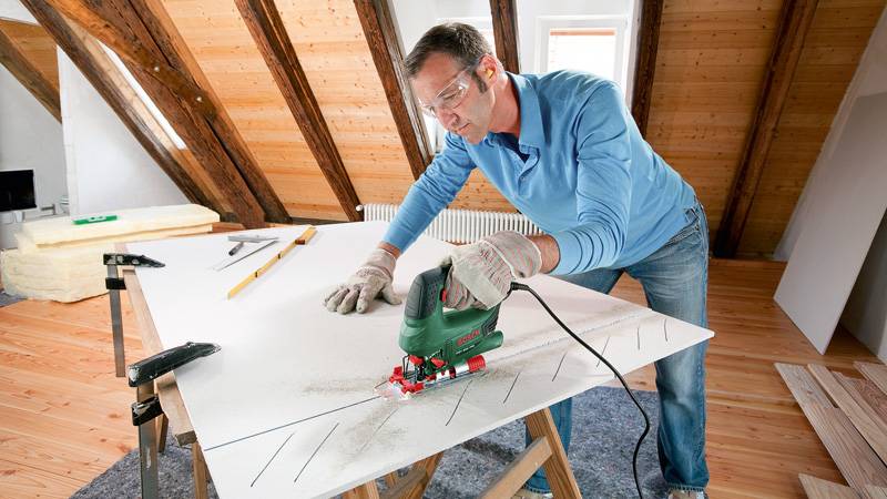 Ein Mann in Schutzausrüstung benutzt eine grüne elektrische Stichsäge, um weißes Plattenmaterial auf einer Werkbank auf einem Dachboden mit freiliegenden Balken zu schneiden.