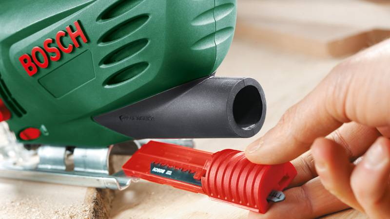 Eine Hand, die einen roten Kunststoff-Staubextraktionadapter in der Nähe des Abgasausgangs einer Bosch-Stichsäge hält, um die Befestigung zur Staubabsaugung zu demonstrieren.