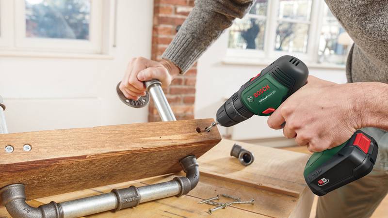Eine Person verwendet einen kabellosen Bohrer, um Schrauben in ein Holzstück zu drehen, das an Metallrohren befestigt ist, und montiert so ein Möbelstück.