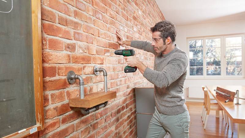 Eine Person nutzt einen Elektroschrauber, um ein Holzregal, das an einer Backsteinmauer befestigt ist, in einem hellen Raum mit einer Tafel und einem großen Fenster zu installieren.