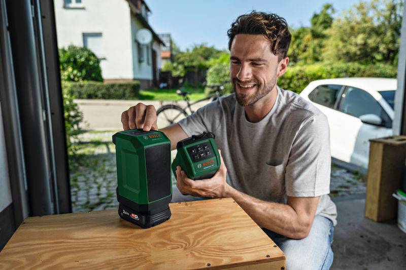 Ein Mann in einem lockeren Hemd lächelt, während er eine grüne Wasserwaage mit Laser auf einem Holztisch in einer Garage aufbaut, mit einem Auto und Fahrrad draußen.