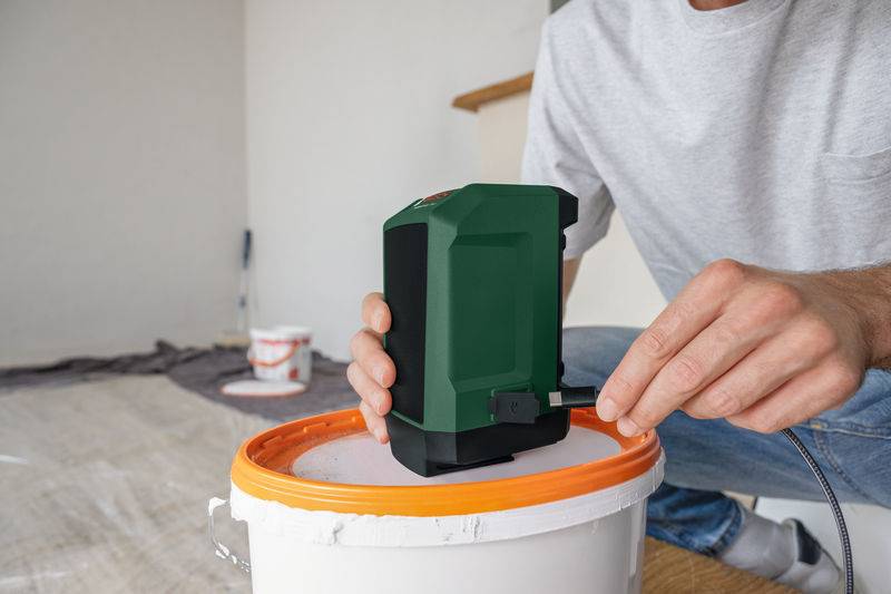 Eine Person verbindet ein Kabel mit einer grünen Laserwasserwage auf einem orange-weiß gestrichenen Farbeimerddeckel, mit Malerbedarfsartikeln im Hintergrund.