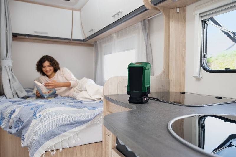 Eine Person liest ein Buch, während sie auf einem Bett in einem Wohnmobil liegt, mit einem tragbaren grünen Gerät auf der Küchentheke im Vordergrund.