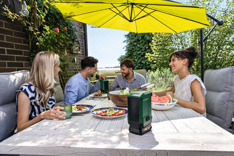 Vier Personen genießen eine Mahlzeit im Freien an einem weißen Tisch mit einem grünen Sonnenschirm, umgeben von Grünflächen und einer Backsteinmauer. Teller mit Speisen stehen auf dem Tisch.
