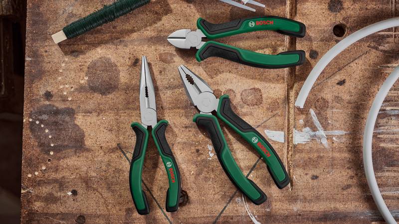 Eine hölzerne Werkbank mit drei grüngriffigen Bosch-Zangen: eine Spitzenzange, eine Seitenschneider und eine Kombizange. Aufgerollter Draht und Schläuche befinden sich in der Nähe.