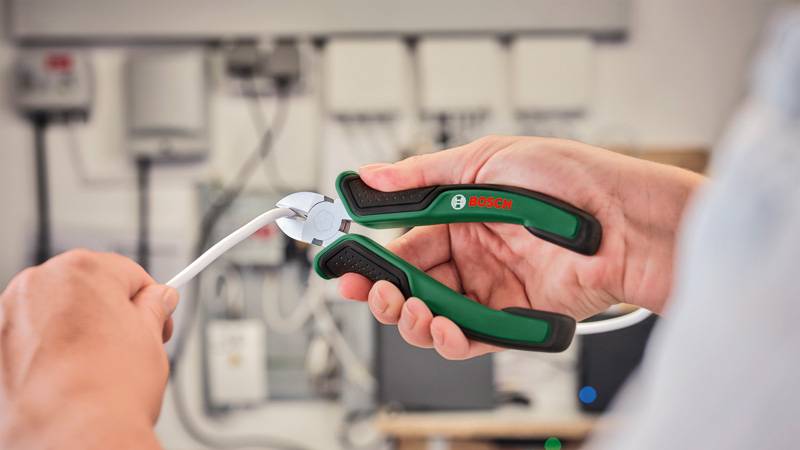 Eine Person verwendet Bosch-Drahtschneider, um ein weißes Kabel in einer Werkstatt zu schneiden, wobei elektronische Geräte im Hintergrund sichtbar sind.