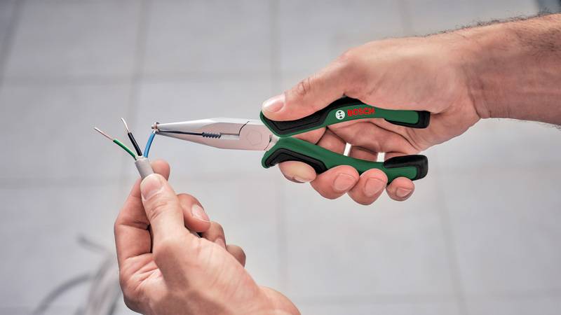 Eine Person verwendet Zangen mit grünem Griff, um die Isolierung von Elektrokabeln zu entfernen und die Kupferlitzen im Inneren freizulegen.