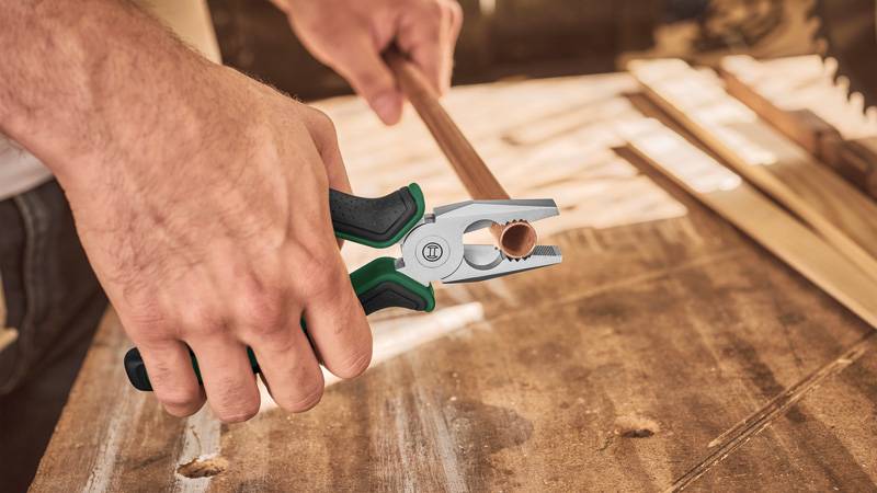 Hände, die mit einer Zange einen Draht auf einem Holztisch abisolieren, Werkzeugbilder im Hintergrund.