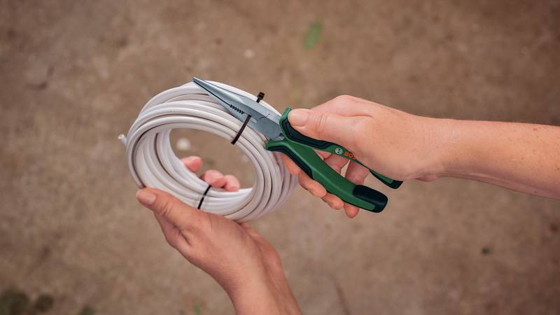 Hände halten Scheren und schneiden einen Kabelbinder auf einer Rolle mit weißem Kabel durch, vor einem Betonhintergrund.