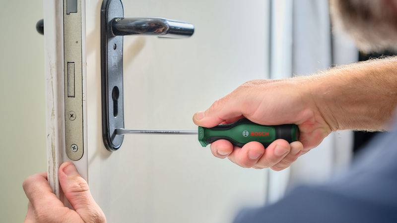 Eine Person benutzt einen Schraubenzieher mit grünem Griff, um ein Türschloss aus einer Holztür zu lösen. Der Fokus liegt auf dem Ausbauvorgang.