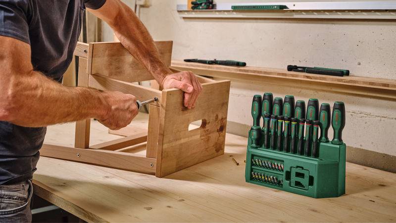 Eine Person montiert eine Holzkiste auf einer Werkbank mit einem Schraubenzieher. Ein grünes Werkzeugsatz mit verschiedenen Schraubenzehern ist in der Nähe sichtbar.