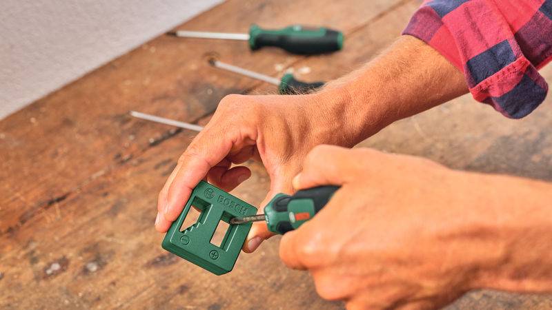 Eine Person benutzt einen Schraubenzieher, um ein grünes Kunststoffobjekt auf einer Holzoberfläche zusammenzusetzen, während andere Werkzeuge im Hintergrund sichtbar sind.