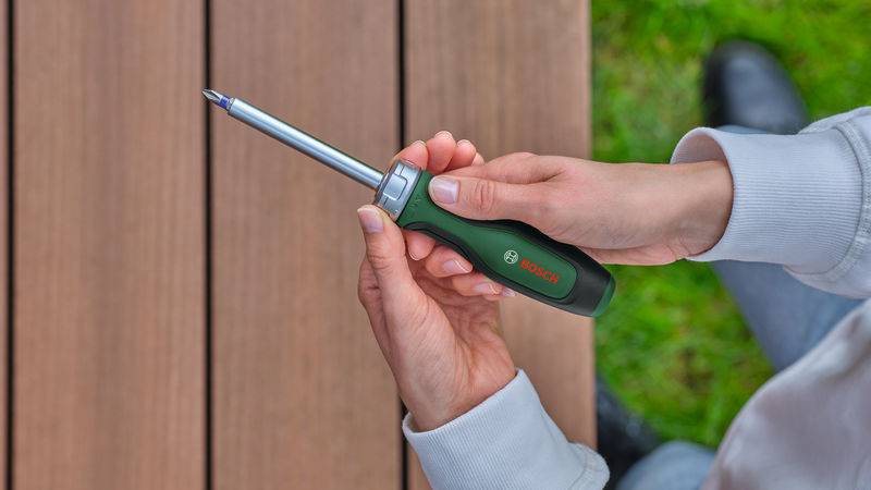 Eine Person, die einen Bosch-Schraubendreher neben einer Holzoberfläche hält, mit einem grasigen Bereich im Hintergrund sichtbar.
