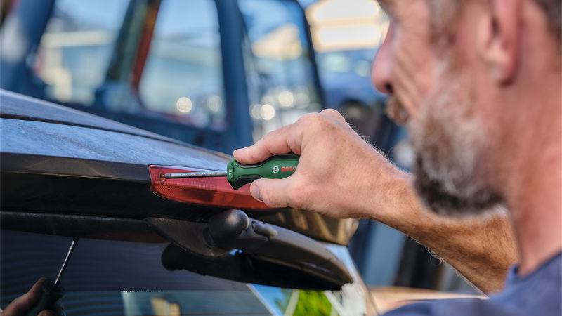 Eine Person nutzt einen Schraubendreher, um eine Schraube auf einem Autodach festzuziehen, wobei sie sich auf den hinteren Bereich konzentriert. Der Hintergrund zeigt unscharfe Fahrzeuge.