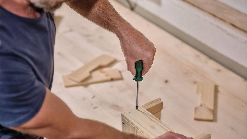 Eine Person nutzt einen Schraubendreher, um ein Möbelstück aus Holz auf einem hellfarbigen Holzboden zusammenzubauen.