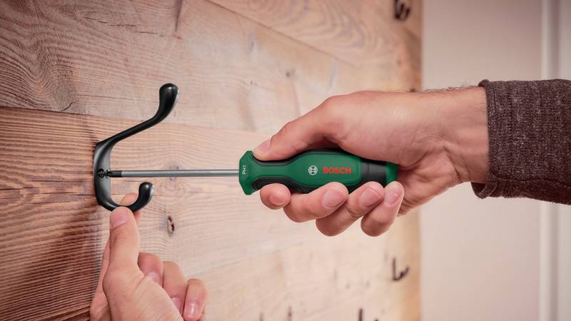 Eine Person verwendet einen Schraubendreher, um einen schwarzen Haken an einer Holzwand zu befestigen und demonstriert damit eine einfache DIY-Aufgabe.