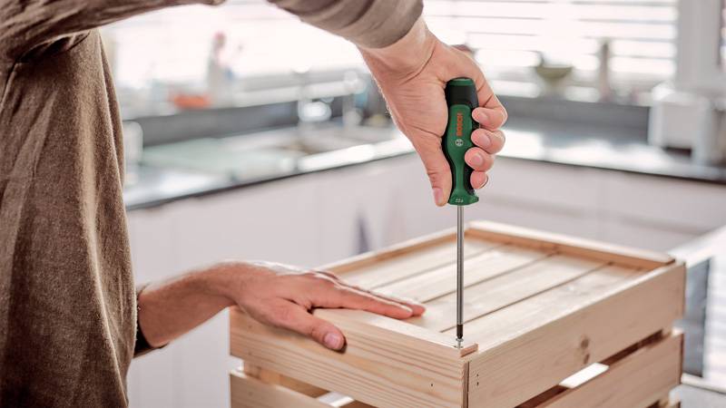 Eine Person schraubt ein Stück Holz mit einem Schraubendreher mit grünem Griff auf eine Holzkiste in einer hellen Küchenumgebung.