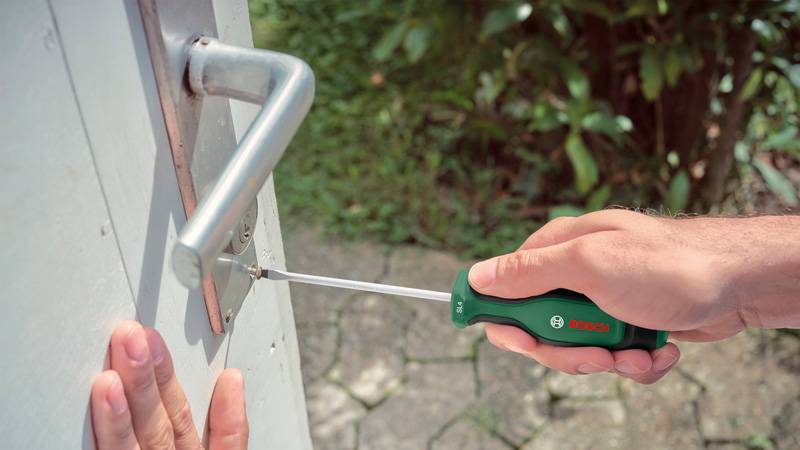 Person, die einen Schraubendreher verwendet, um ein Scharnier an einer Tür anzuziehen oder zu lockern. Die Szene spielt sich im Freien ab und deutet auf laufende Wartungs- oder Reparaturarbeiten hin.