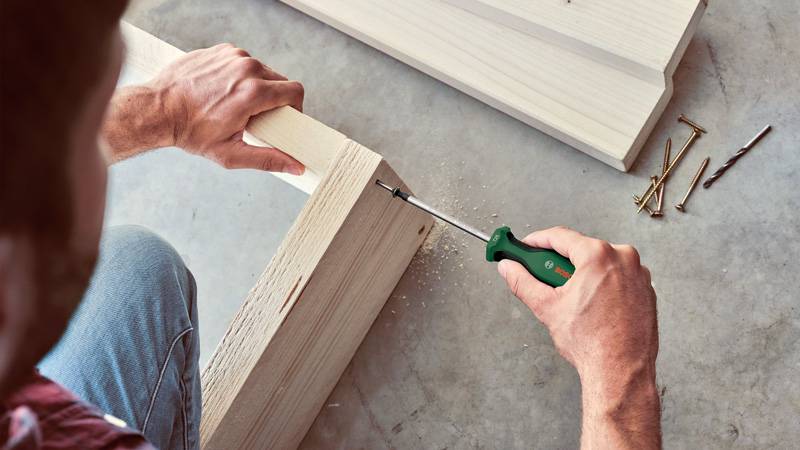 Eine Person verwendet einen grünen Schraubendreher, um Holzteile auf einem Betonboden zusammenzusetzen. Mehrere Schrauben und Nägel sind in der Nähe verstreut.