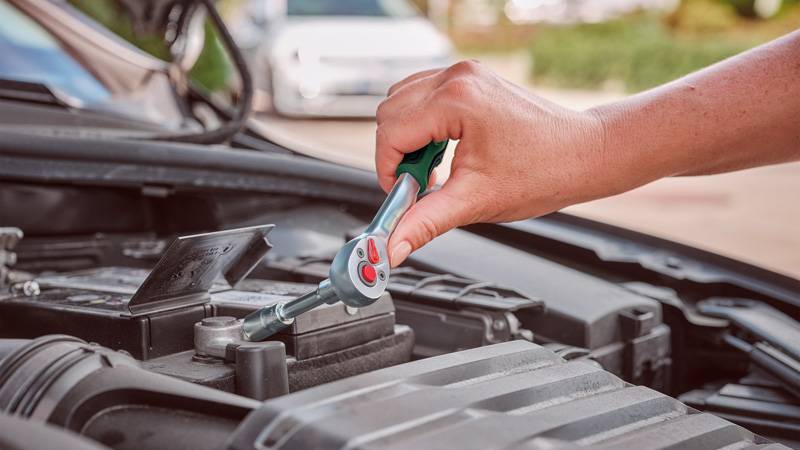 Eine Person nutzt einen Schraubenschlüssel, um eine Schraube an einem Automotor einzustellen, was auf Wartungs- oder Reparaturarbeiten hindeutet. Im Hintergrund ist ein geparktes Auto zu sehen.