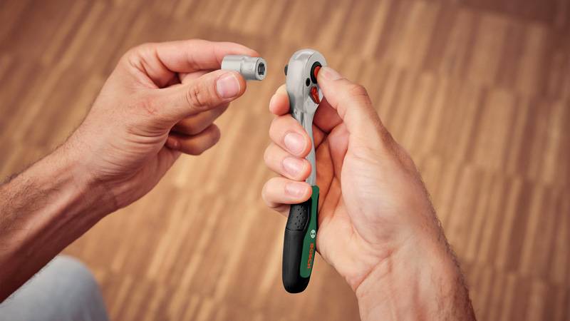 Eine Person hält einen Ratschenschlüssel in der rechten Hand und einen Steckaufsatz in der linken Hand über einem Holzboden, bereit zum Zusammenbau.